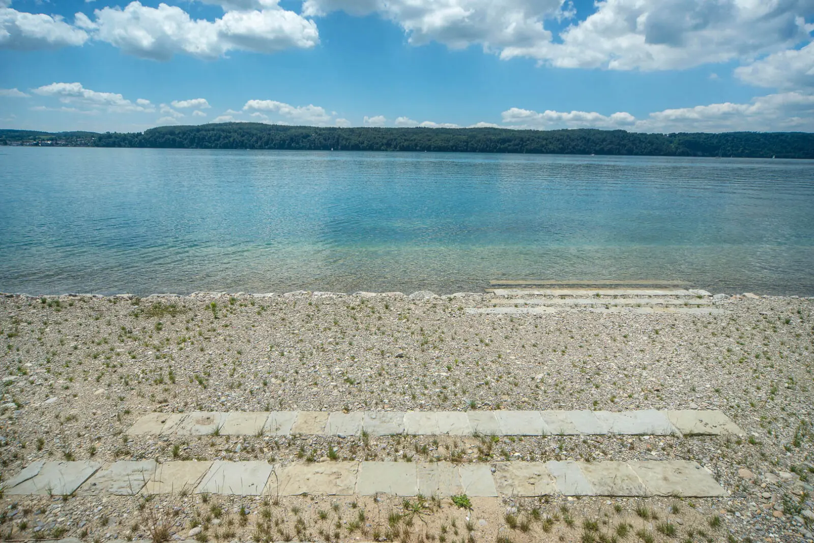 Natural lakeside edge featuring a pebble beach, stone access steps, and clear water, creating a simple and inviting connection between landscape and shoreline.